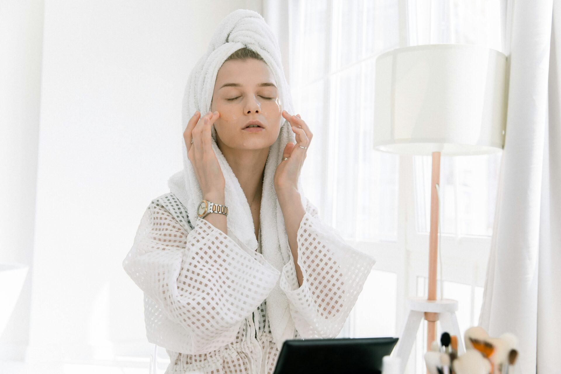 Adult woman in bathrobe doing skincare routine with head towel and under-eye patches indoors.